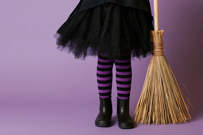 Child's legs in witch costume with straw broom on purple background