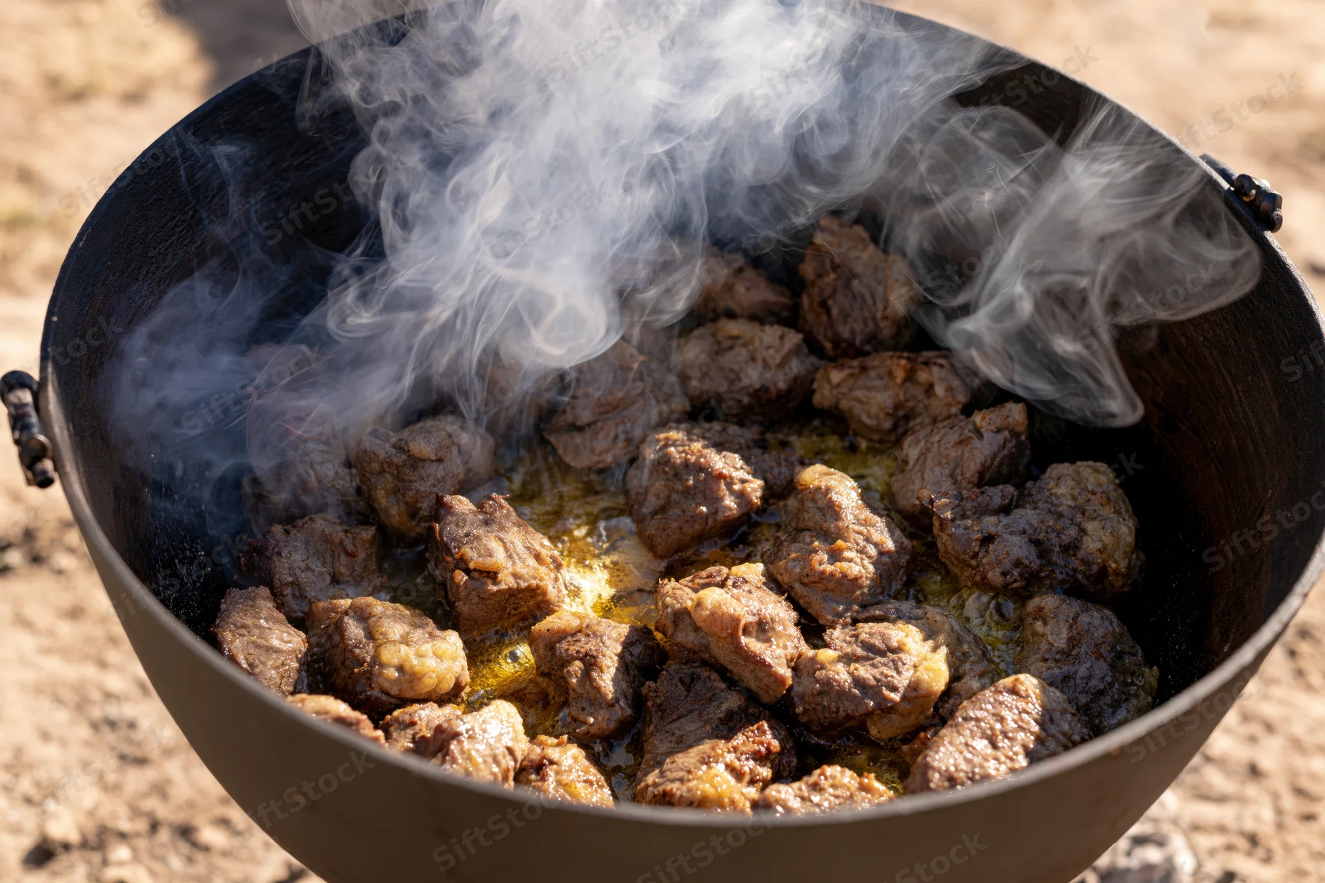 Chunks of seared beef cooking in a blackened cast iron pot over an open fire outdoors