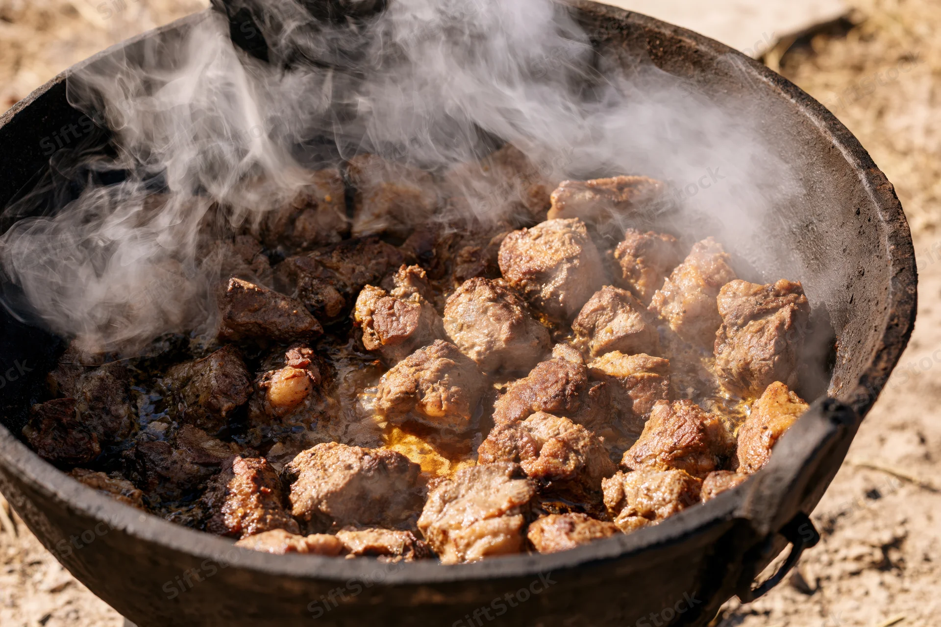 Chunks of seared meat cooking in a large blackened cast iron pot outdoors
