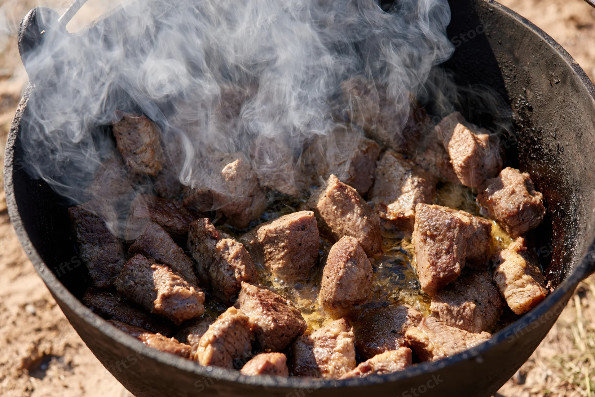 Chunks of seared beef cooking in a blackened cast iron pot over an open fire