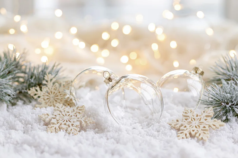 Clear glass christmas ornaments resting in artificial snow with pine branches and snowflakes, warm lights in background