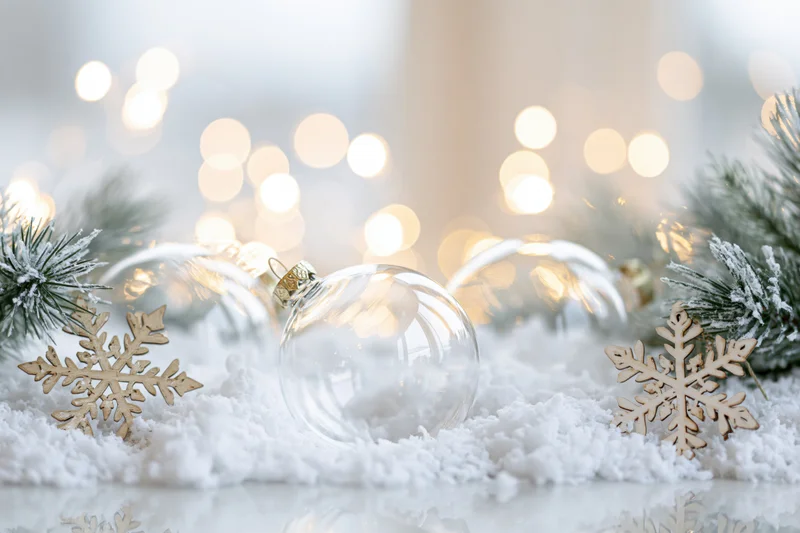 Clear glass christmas ornaments resting in artificial snow with wooden snowflakes and pine branches, illuminated by warm bokeh lights