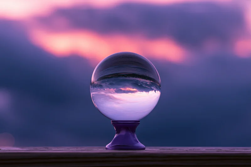 Crystal ball on purple pedestal reflecting sunset sky on wooden surface