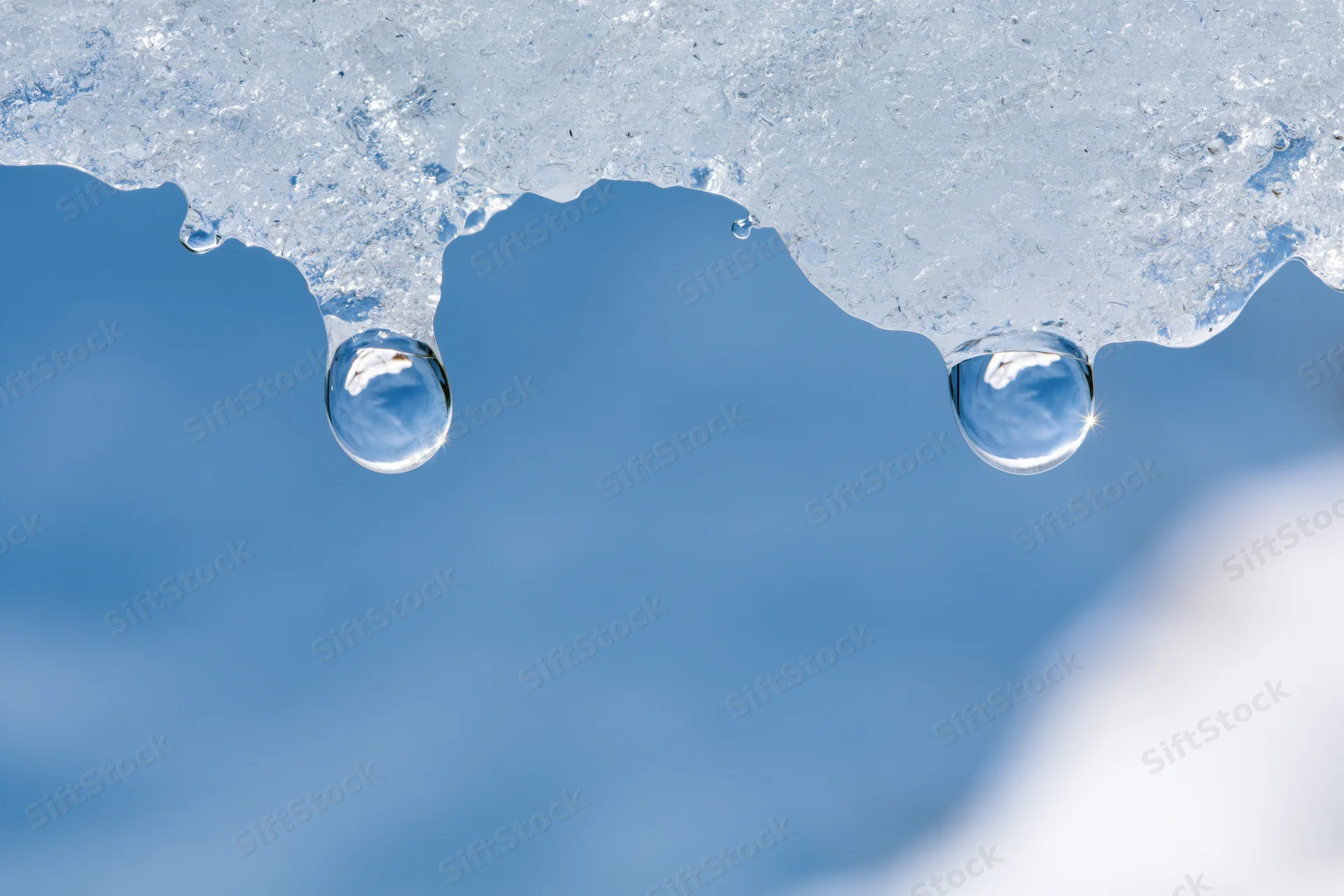 Two water droplets hanging from melting ice against a blue sky
