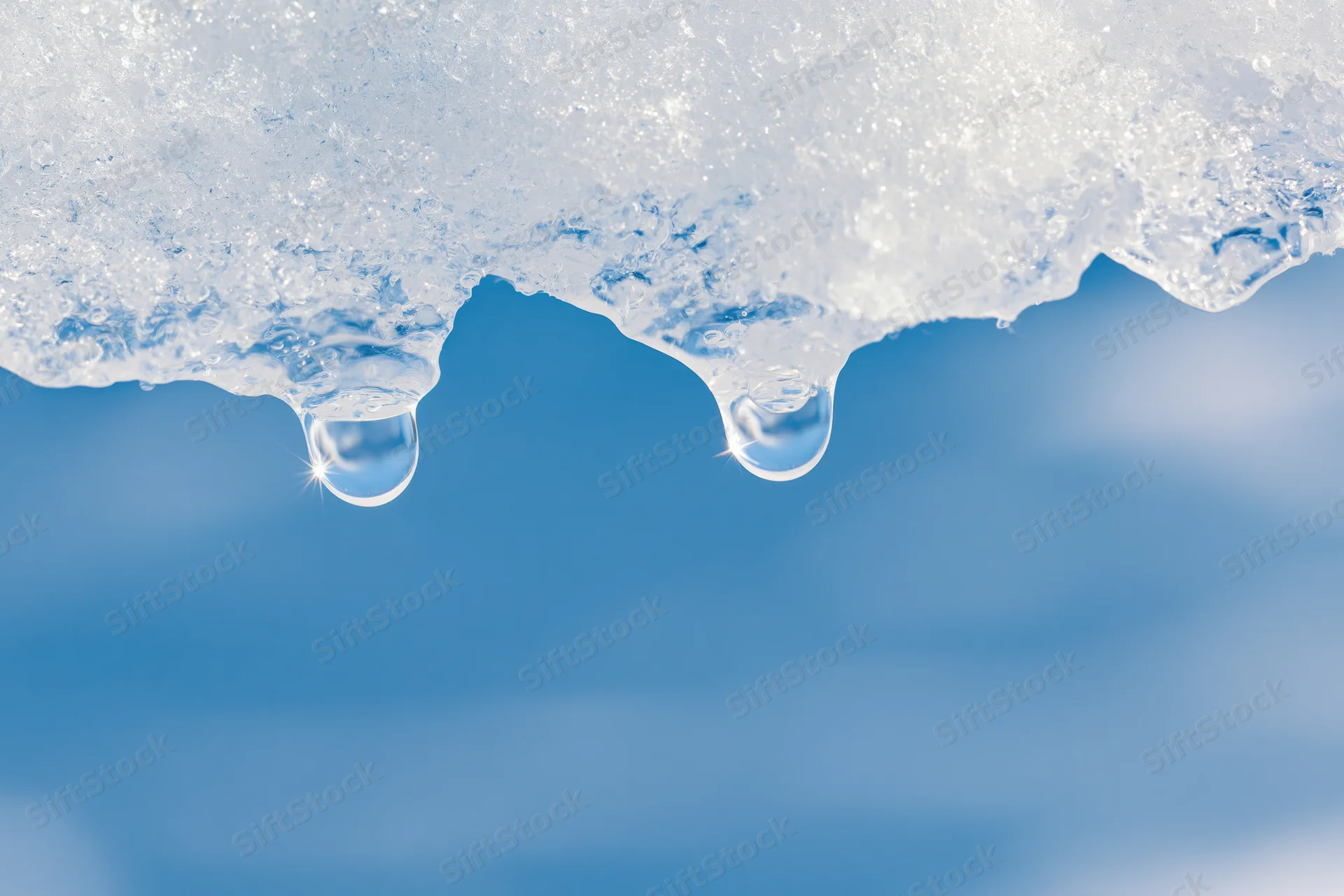 Two water droplets hanging from melting ice against a blue sky background