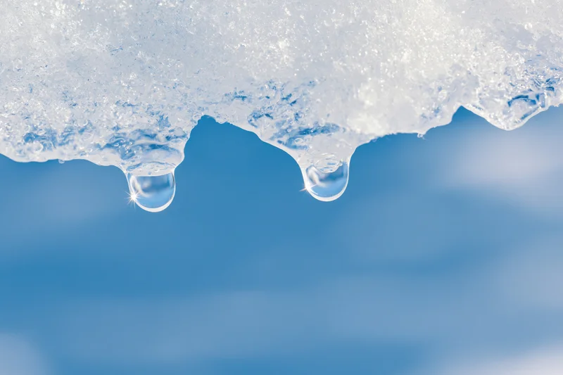 Two water droplets hanging from melting ice against a blue sky background