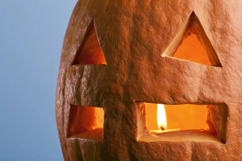 Close-up of a carved pumpkin with triangular eyes and rectangular mouth illuminated by a candle inside against a blue background