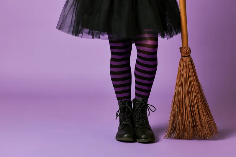 Child in witch costume standing beside broom on purple background