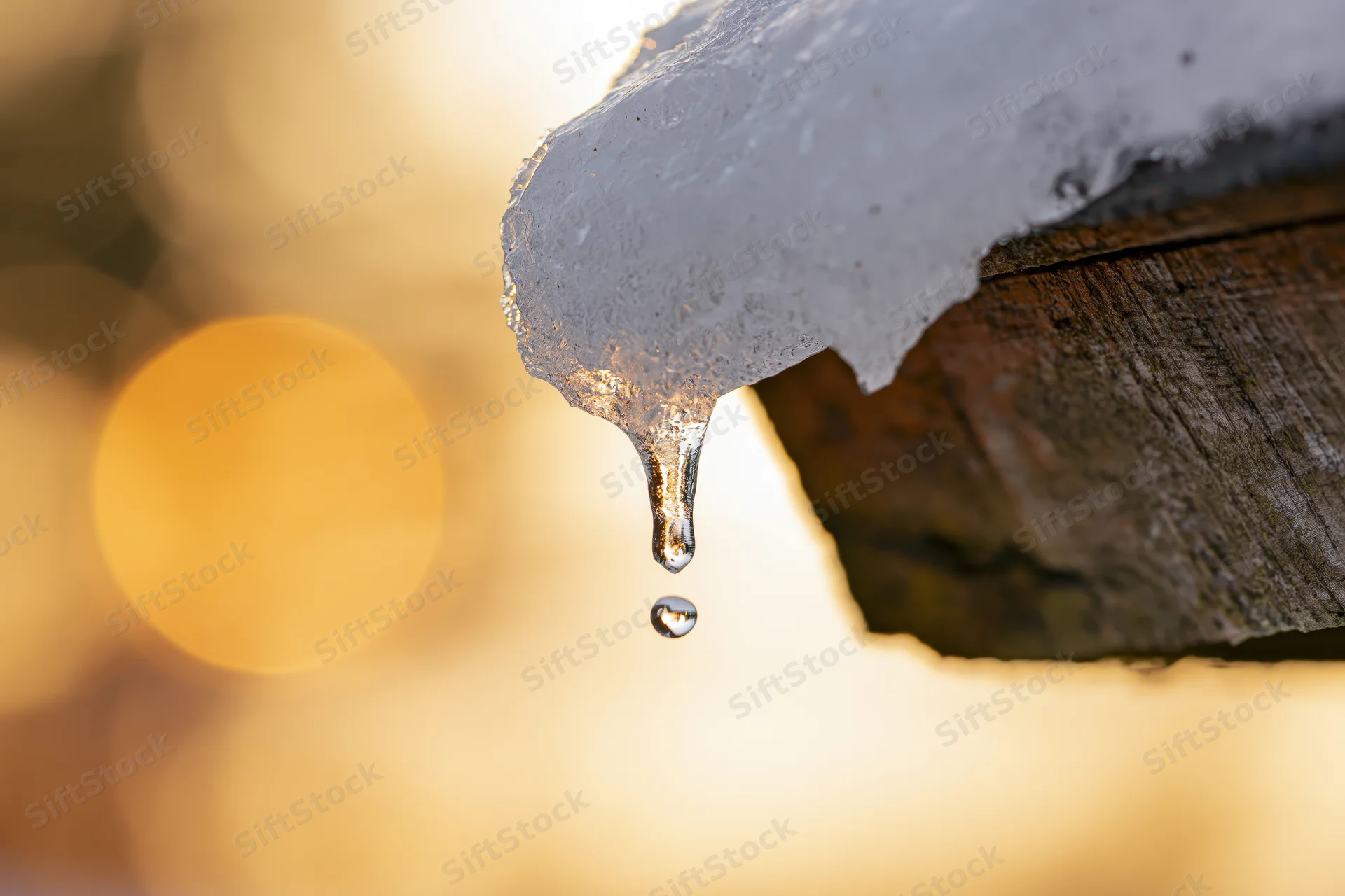 Melting ice dripping from a wooden roof eave with golden bokeh background