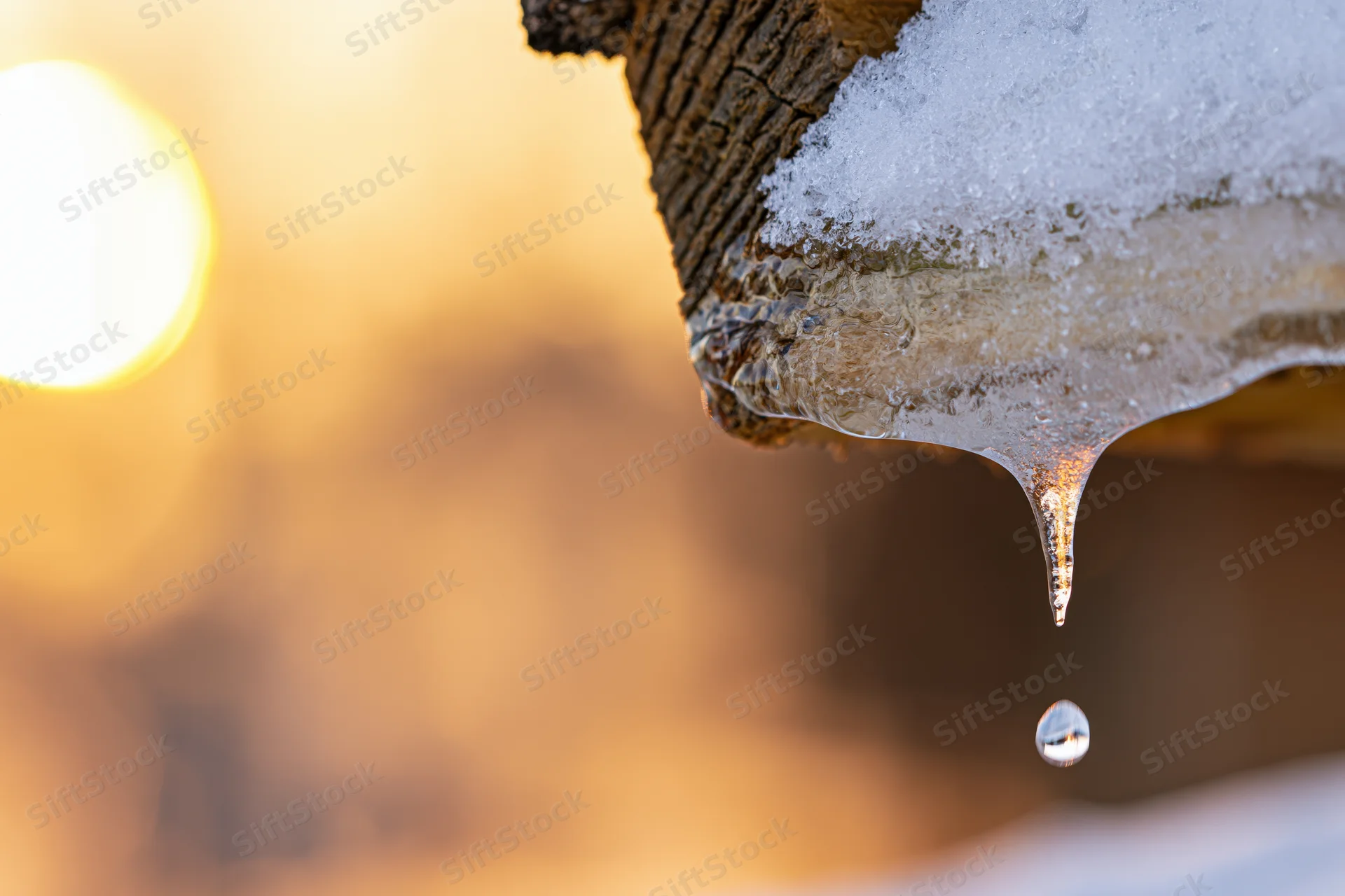 Icicle dripping from a wooden roof eave in winter with golden bokeh light in the background