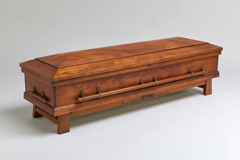 Closed wooden coffin with brass handles resting on matching wooden stand against white background