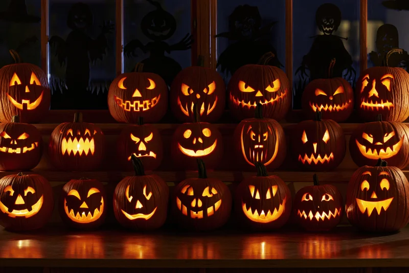 Carved jack-o-lanterns glowing on a wooden table in front of a window with witch and ghost silhouettes