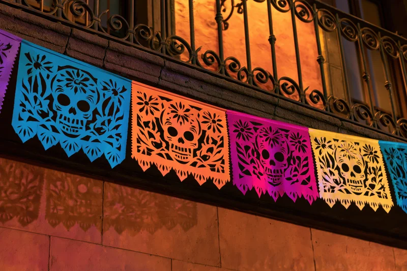 Colorful papel picado banners with skull and floral designs strung beneath a wrought-iron balcony at night