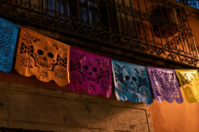 Colorful papel picado banners with skull designs strung beneath a wrought-iron balcony at night