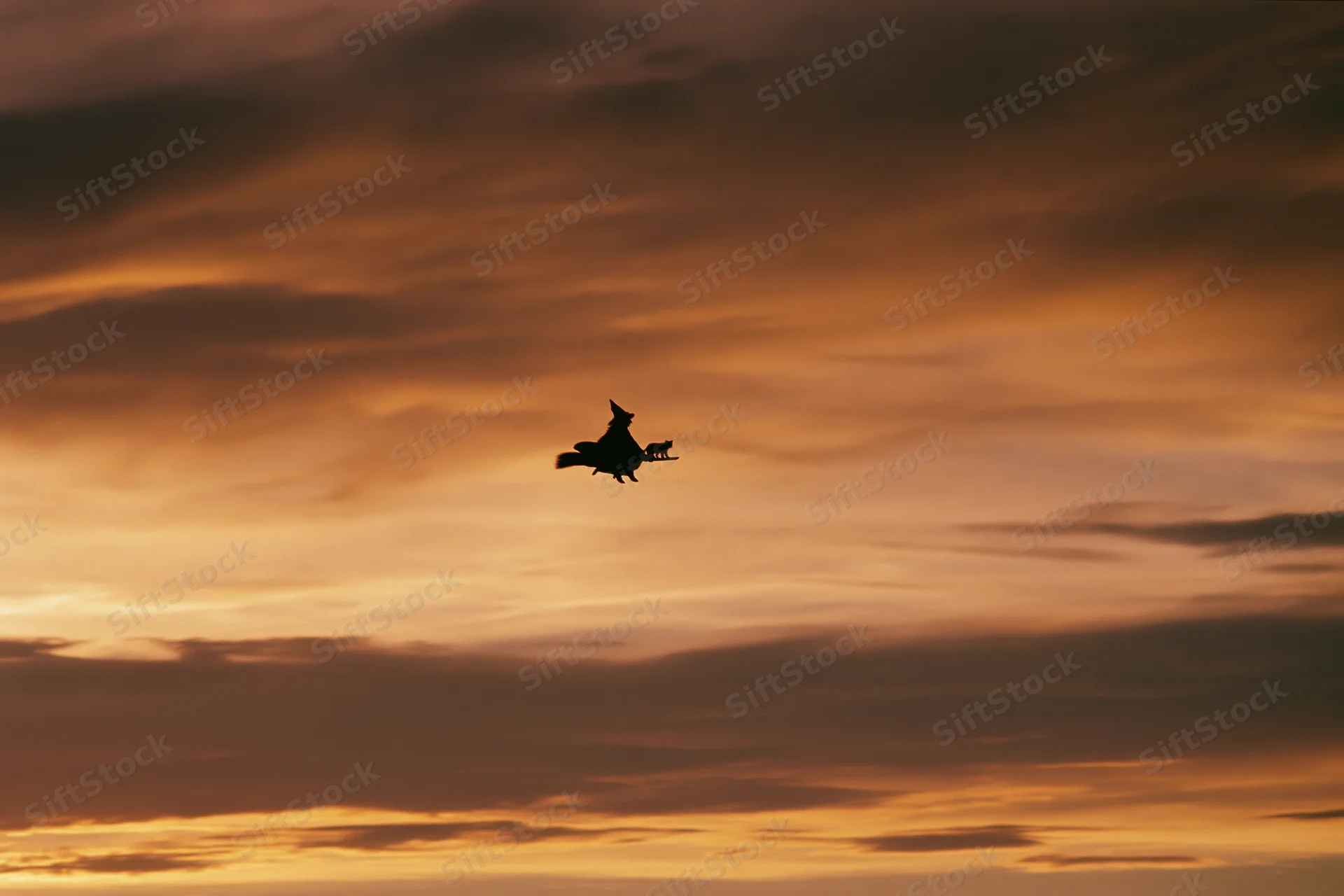 Silhouette of a witch riding a broomstick across a cloudy sunset sky