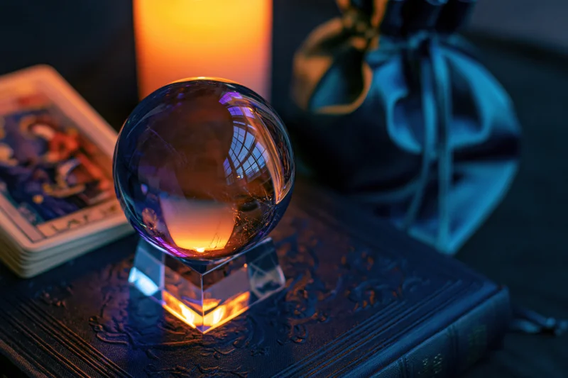 Crystal ball on acrylic stand resting on dark embossed book beside tarot cards and candle