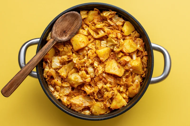 Braised cabbage and potato stew in a cast iron pot with wooden spoon on yellow background