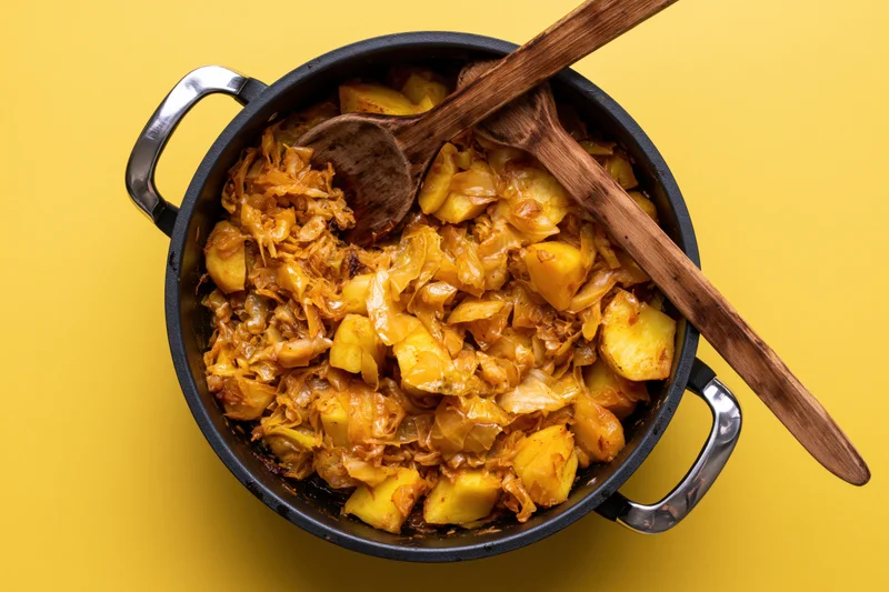 Braised cabbage and potato stew in a cast iron pot with wooden spoons on yellow background