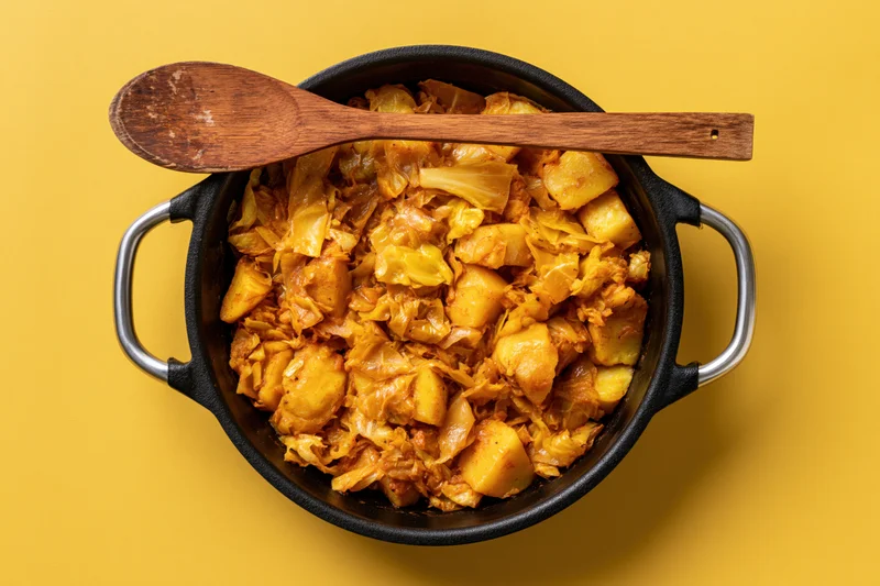 Braised cabbage and potato stew in a cast iron pot with wooden spoon on yellow background