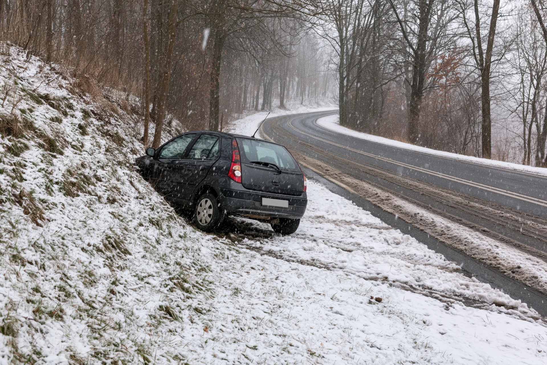 Black compact car sliding off snowy rural road into ditch beside forested hillside