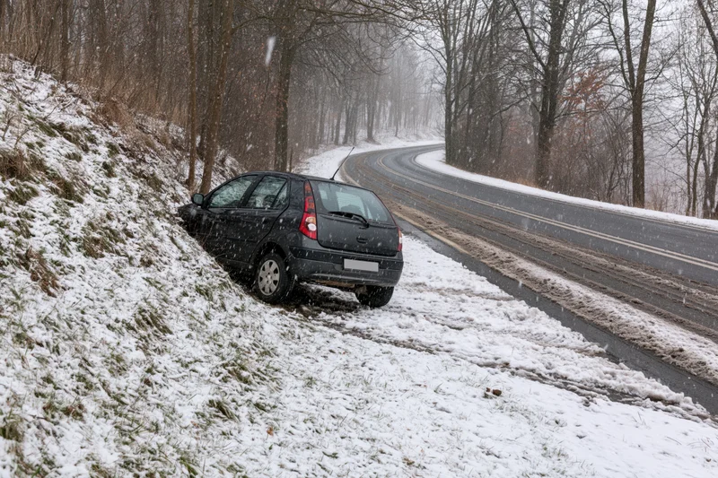Black compact car sliding off snowy rural road into ditch beside forested hillside