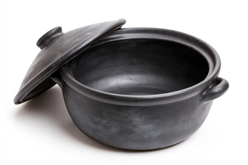 Black clay cooking pot with lid resting against it on white background