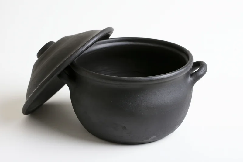 Black clay cooking pot with lid resting against it on white background