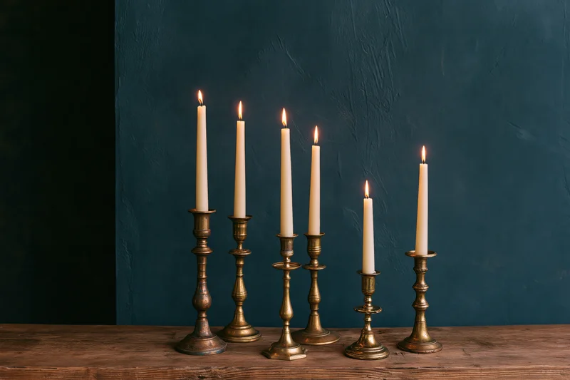 Five lit taper candles in mismatched antique brass candlesticks arranged on a wooden table against a dark blue textured wall