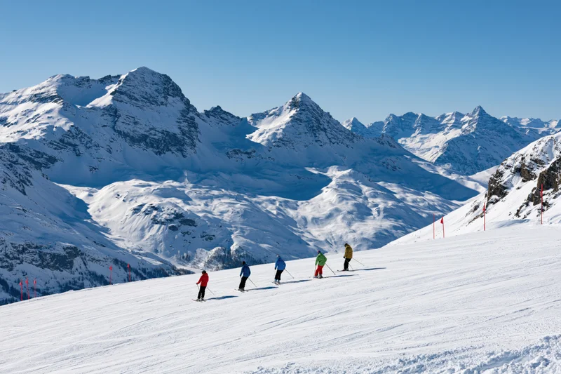 Five skiers descending a groomed alpine slope under clear blue sky
