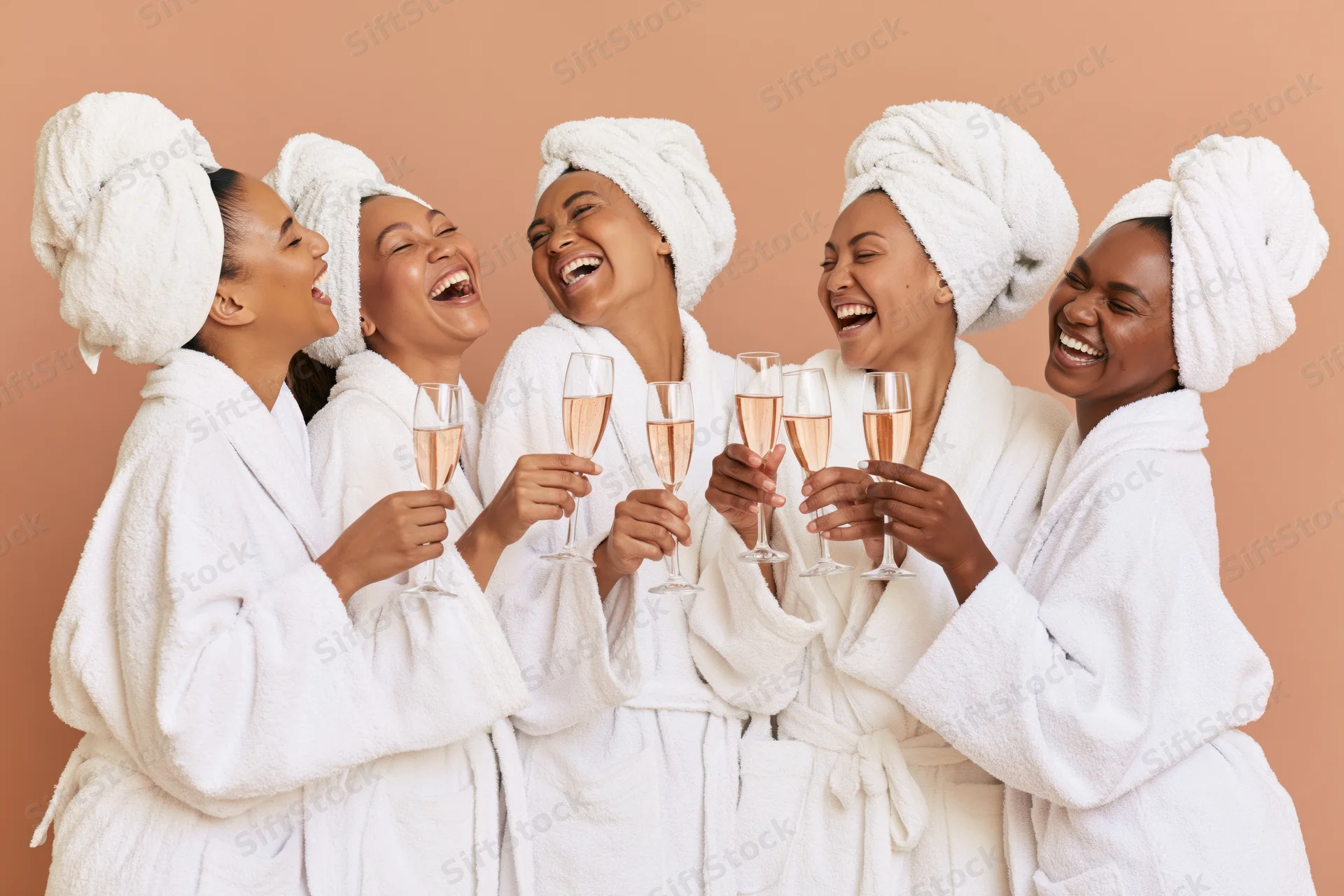 Five women in white bathrobes and towels laughing while toasting with champagne glasses
