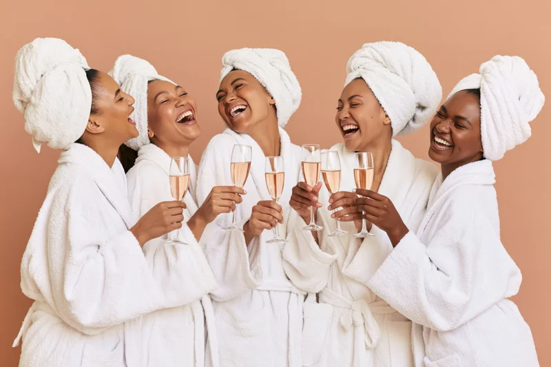 Five women in white bathrobes and towels laughing while toasting with champagne glasses