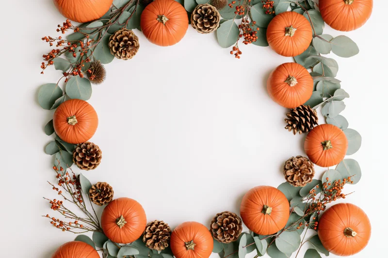Circular arrangement of orange pumpkins, pinecones, eucalyptus, and berries forming a wreath on white surface with central copyspace