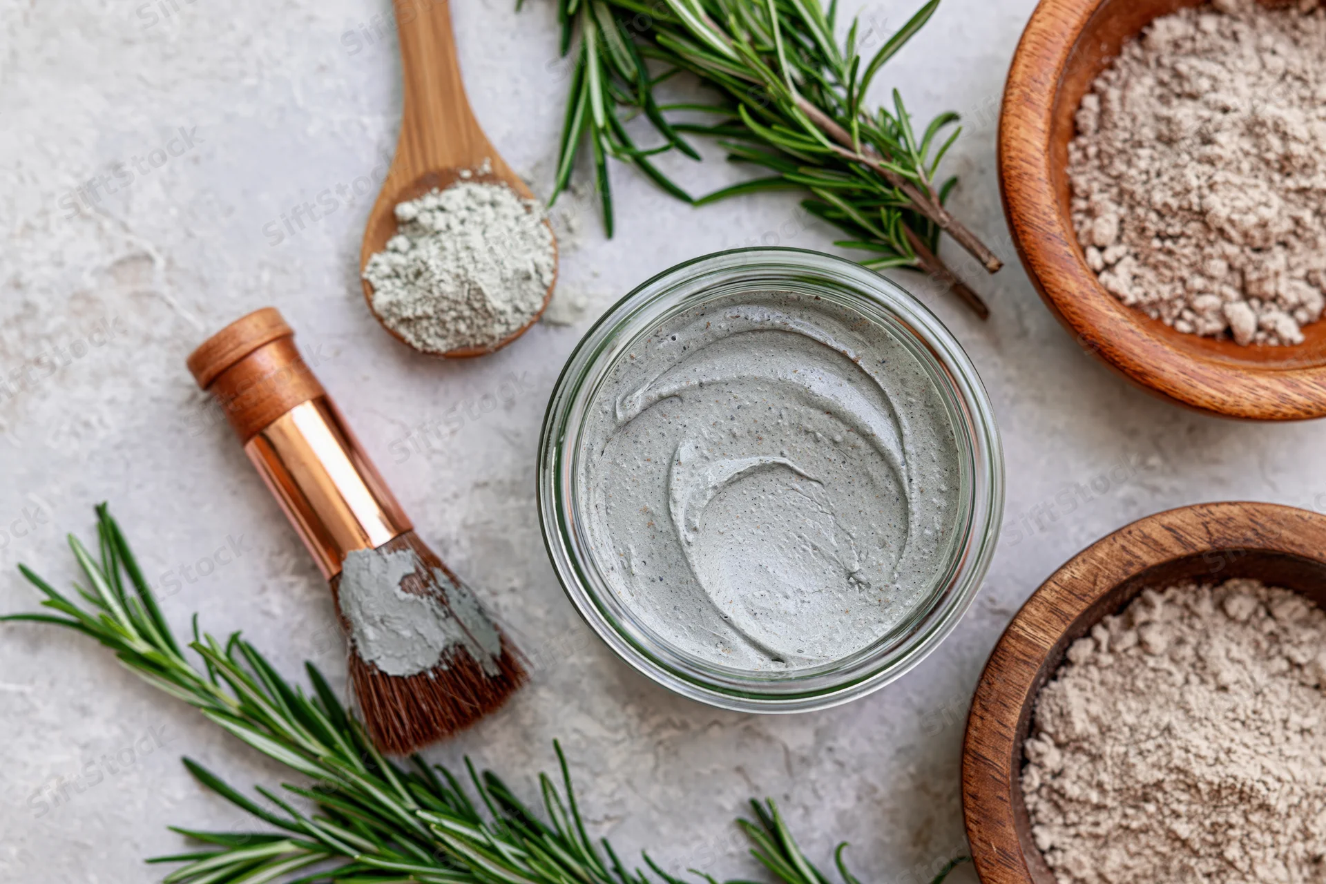 Glass jar with gray clay face mask surrounded by rosemary sprigs, wooden bowls of powder, and a brush on a light surface