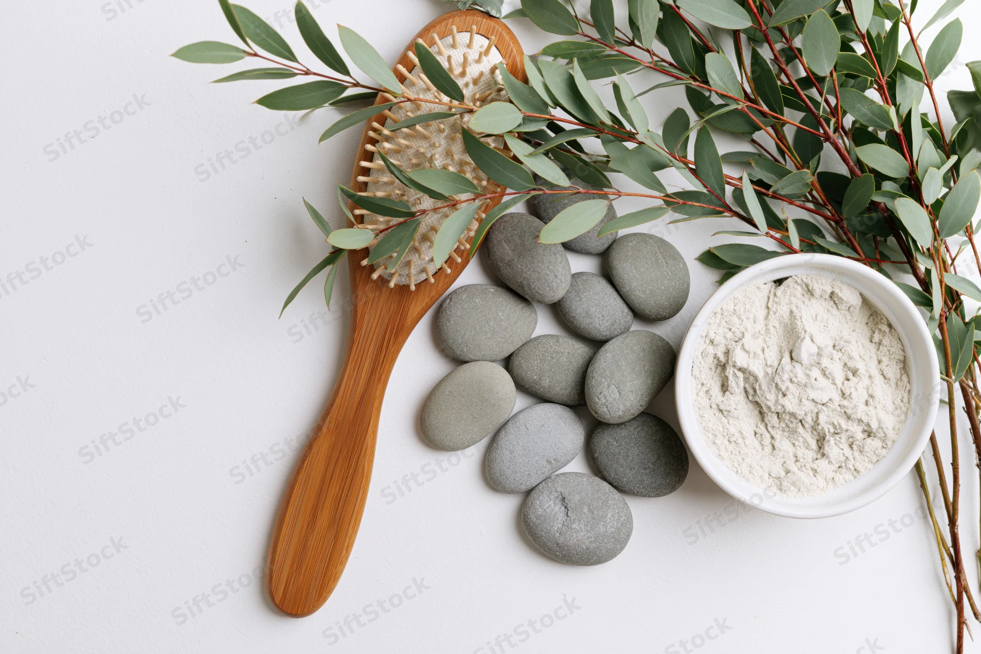 Wooden hairbrush with eucalyptus branches, smooth stones and white powder in ceramic bowl on white surface
