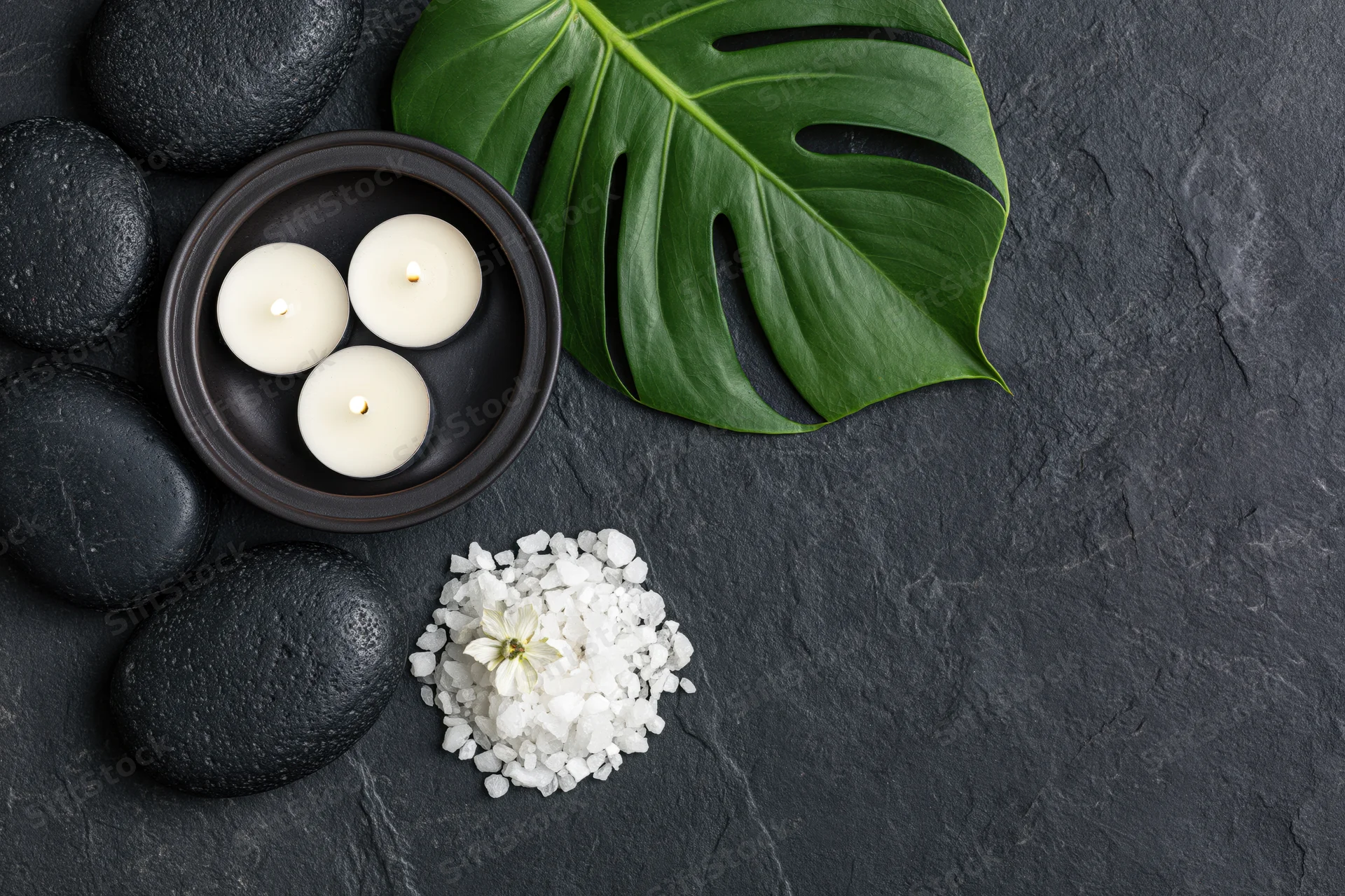 Black basalt stones, lit candles, monstera leaf and white crystals arranged on dark slate surface