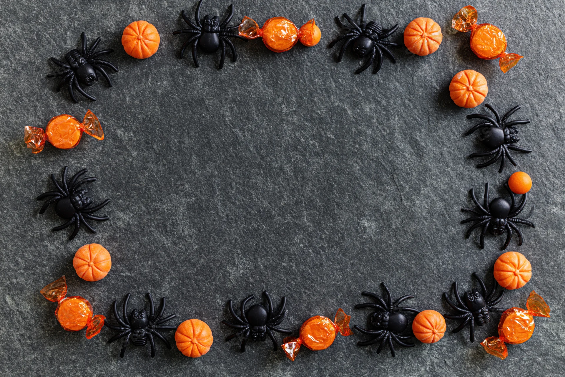 Plastic spiders and orange candies arranged in a frame on dark slate surface