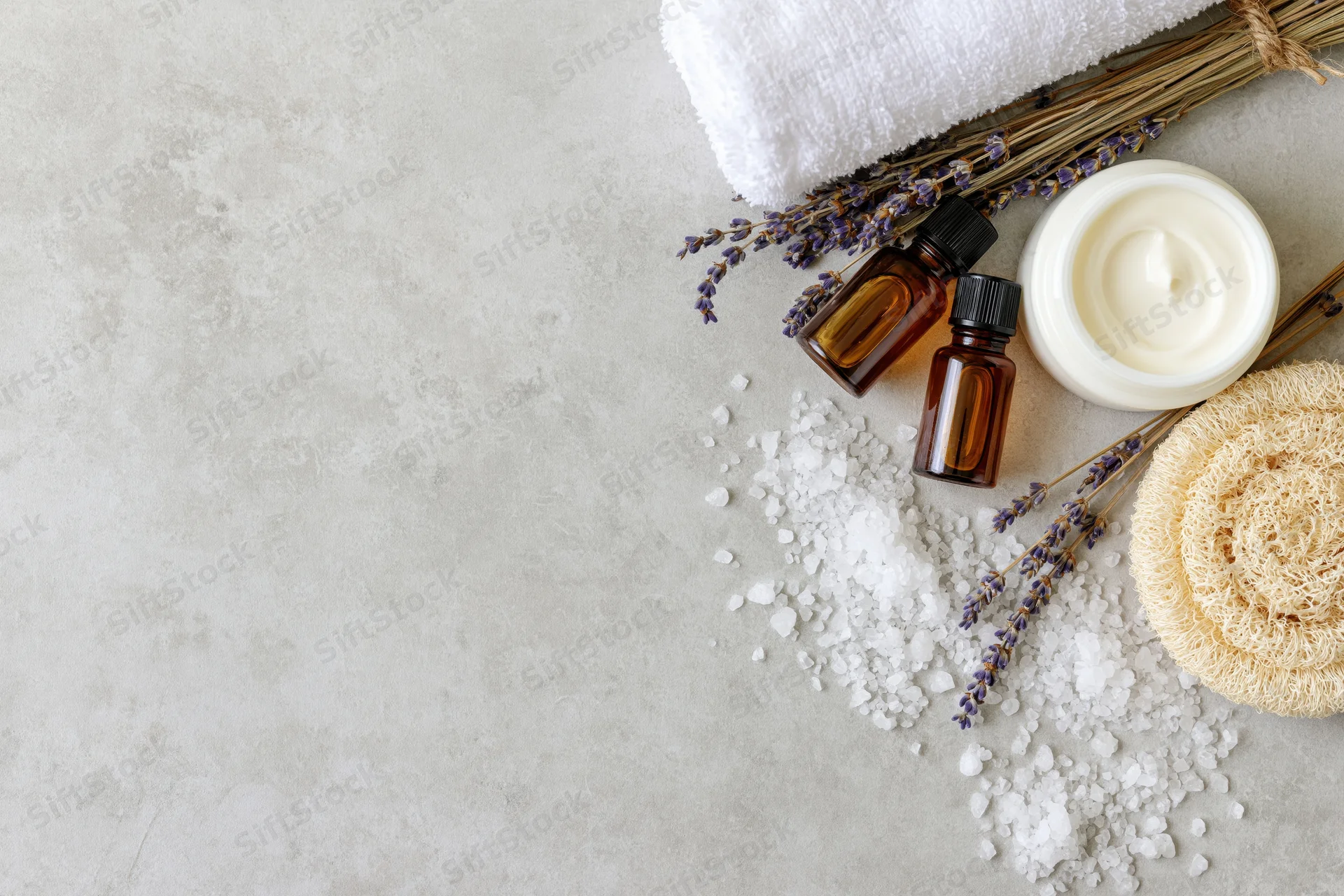 Skincare and spa products arranged with lavender, bath salt, cream, towel and loofah on a textured surface