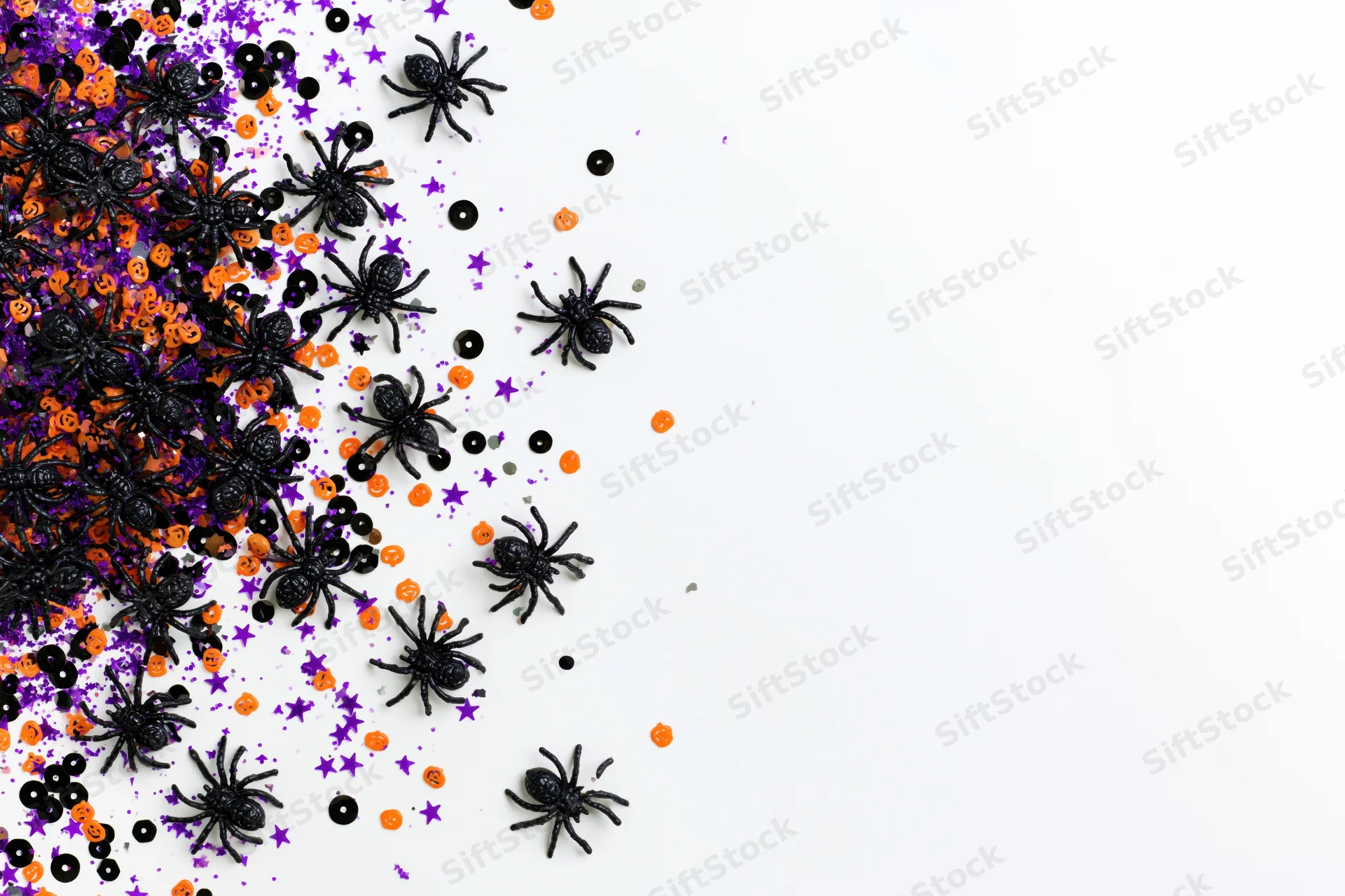 Small black plastic toy spiders scattered across a white surface with orange and purple confetti for halloween decoration