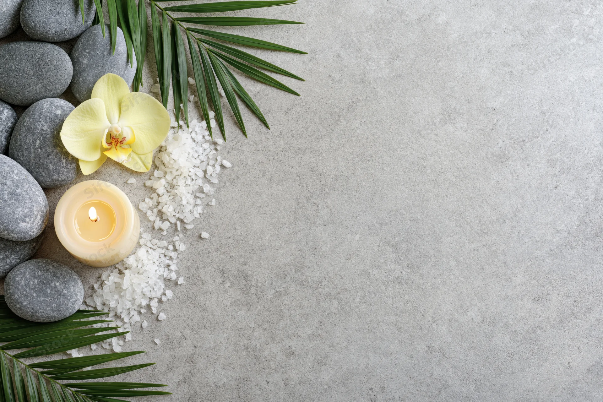 Smooth grey spa stones, lit cream candle, yellow orchid, palm leaves, and bath salt arranged on concrete surface in flat lay with copyspace