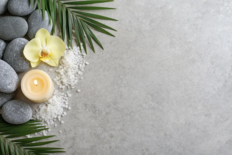 Smooth grey spa stones, lit cream candle, yellow orchid, palm leaves, and bath salt arranged on concrete surface in flat lay with copyspace