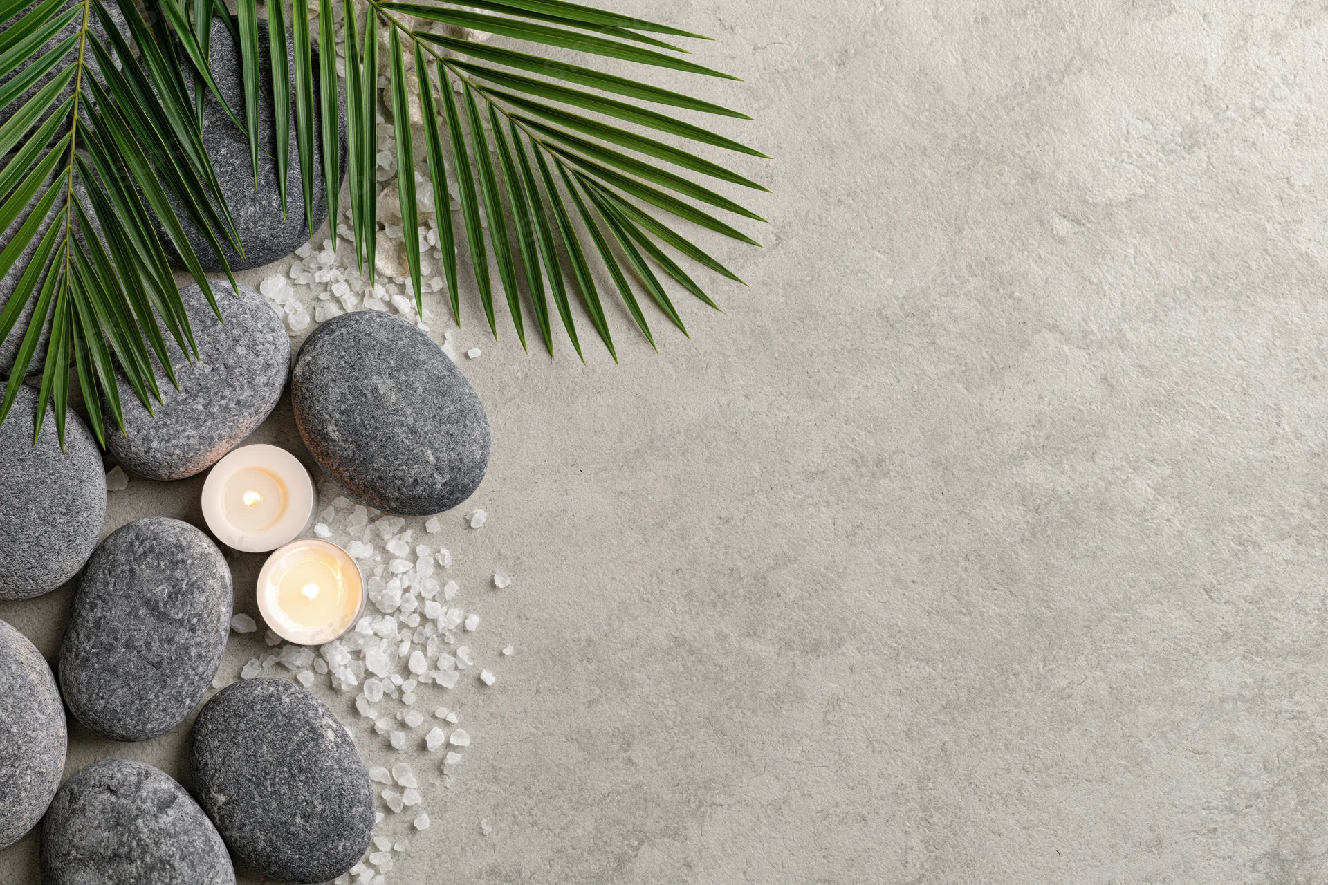 Smooth grey spa stones and lit candle with palm leaves arranged on concrete surface