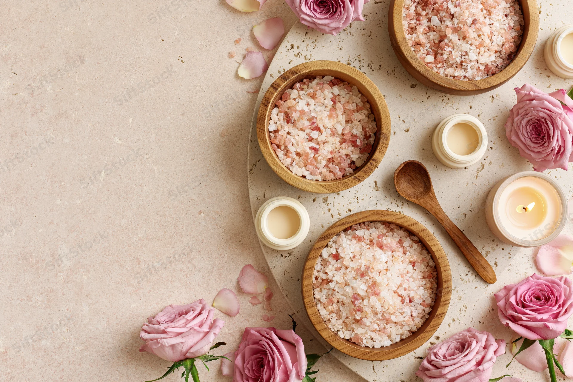 Spa and skincare items including bath salts, roses, candles, and wooden bowls arranged on a pale blush surface with copyspace