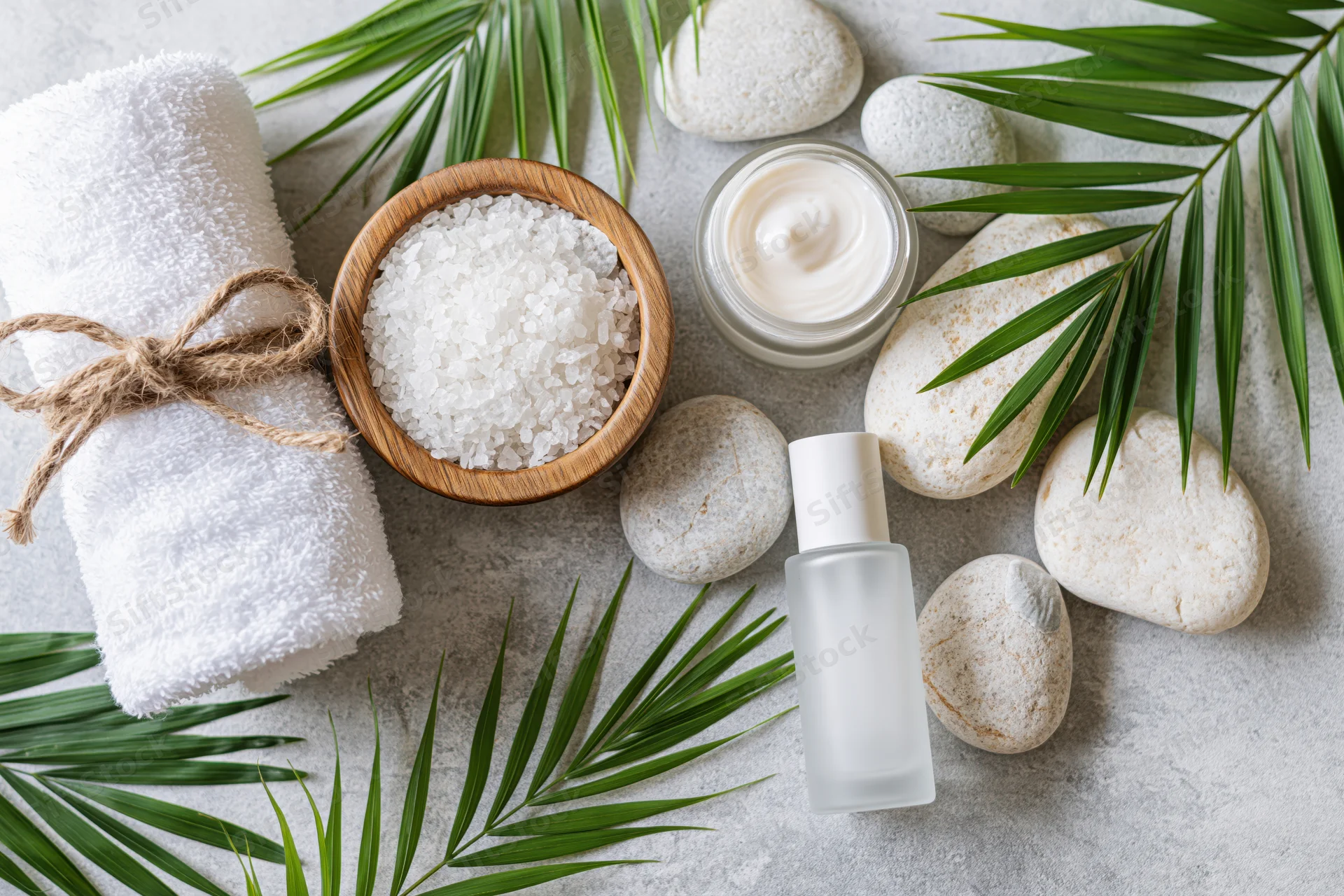 Spa products including cream, serum, salt and towel arranged with palm leaves and stones on a light surface