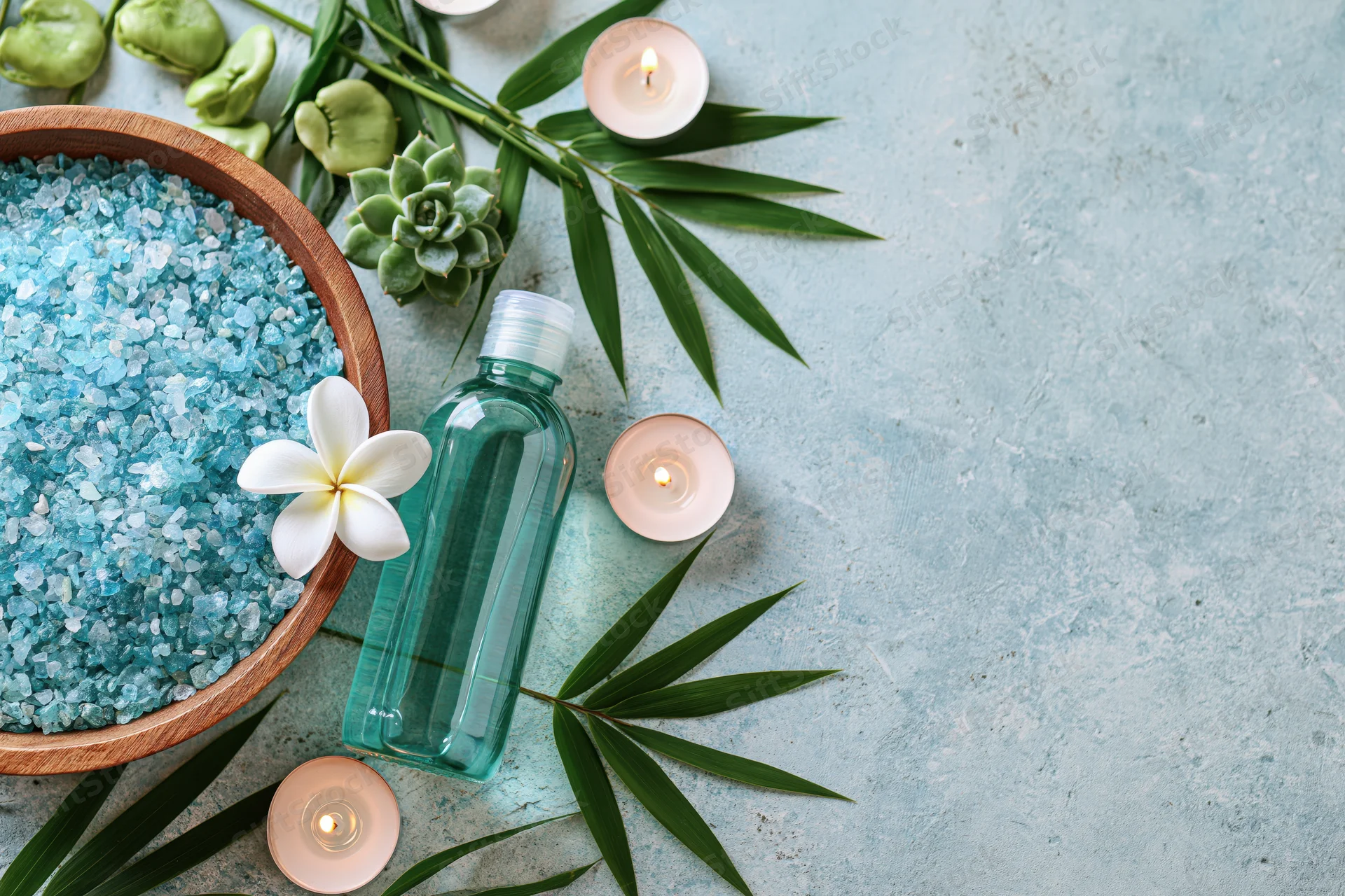 Spa wellness products arranged on pale blue textured surface with candles, bamboo leaves, succulents and bath salts