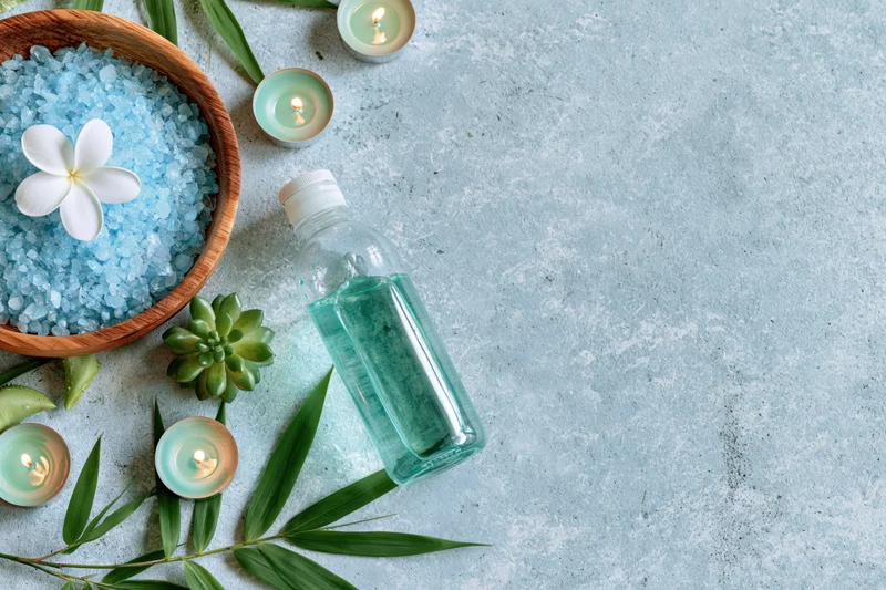 Spa wellness products arranged on pale blue textured surface with candles, salt, plant leaves and bottle of oil