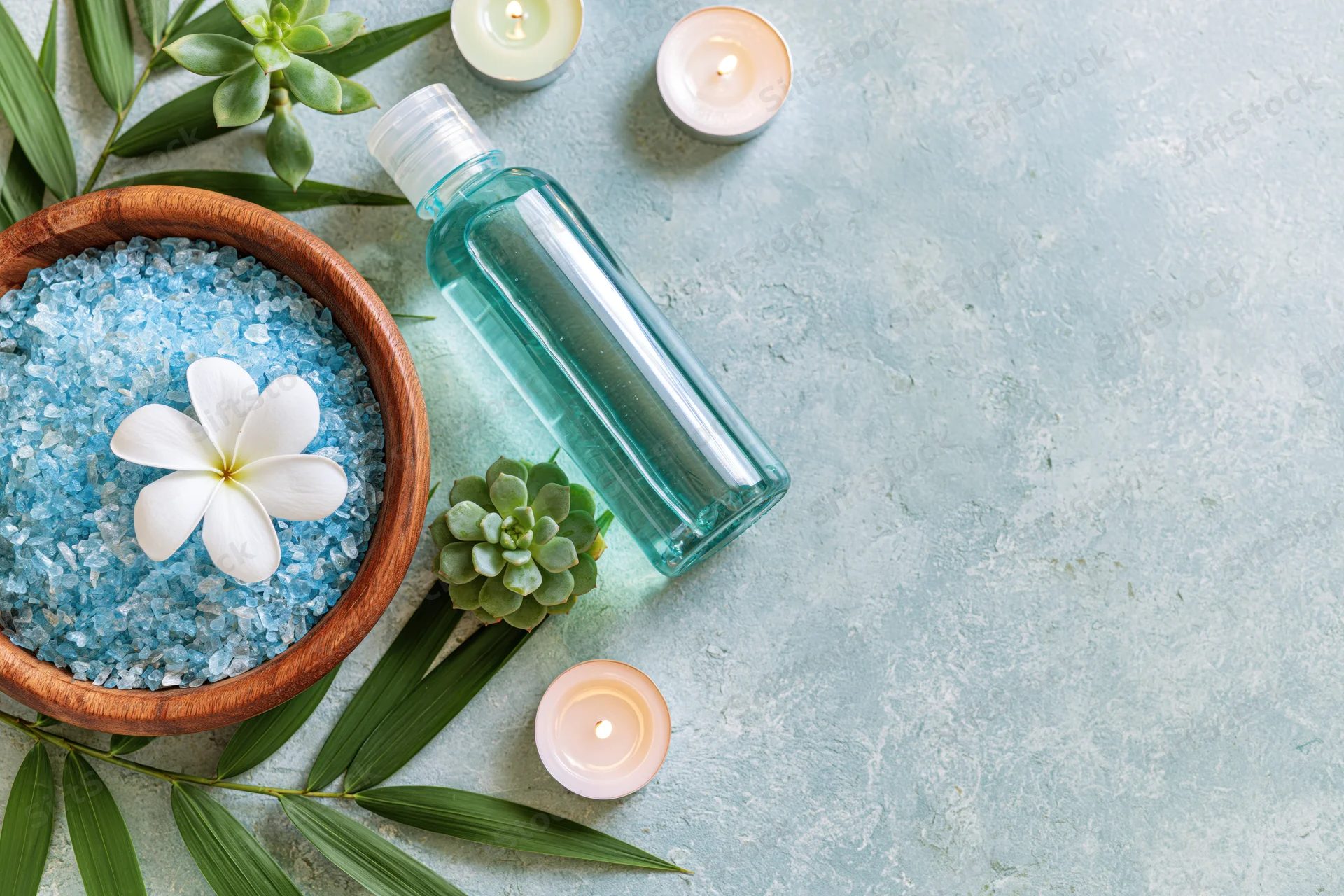 Spa wellness products arranged in flat lay with bath salts, essential oil, candles, succulents and bamboo leaves on textured blue surface