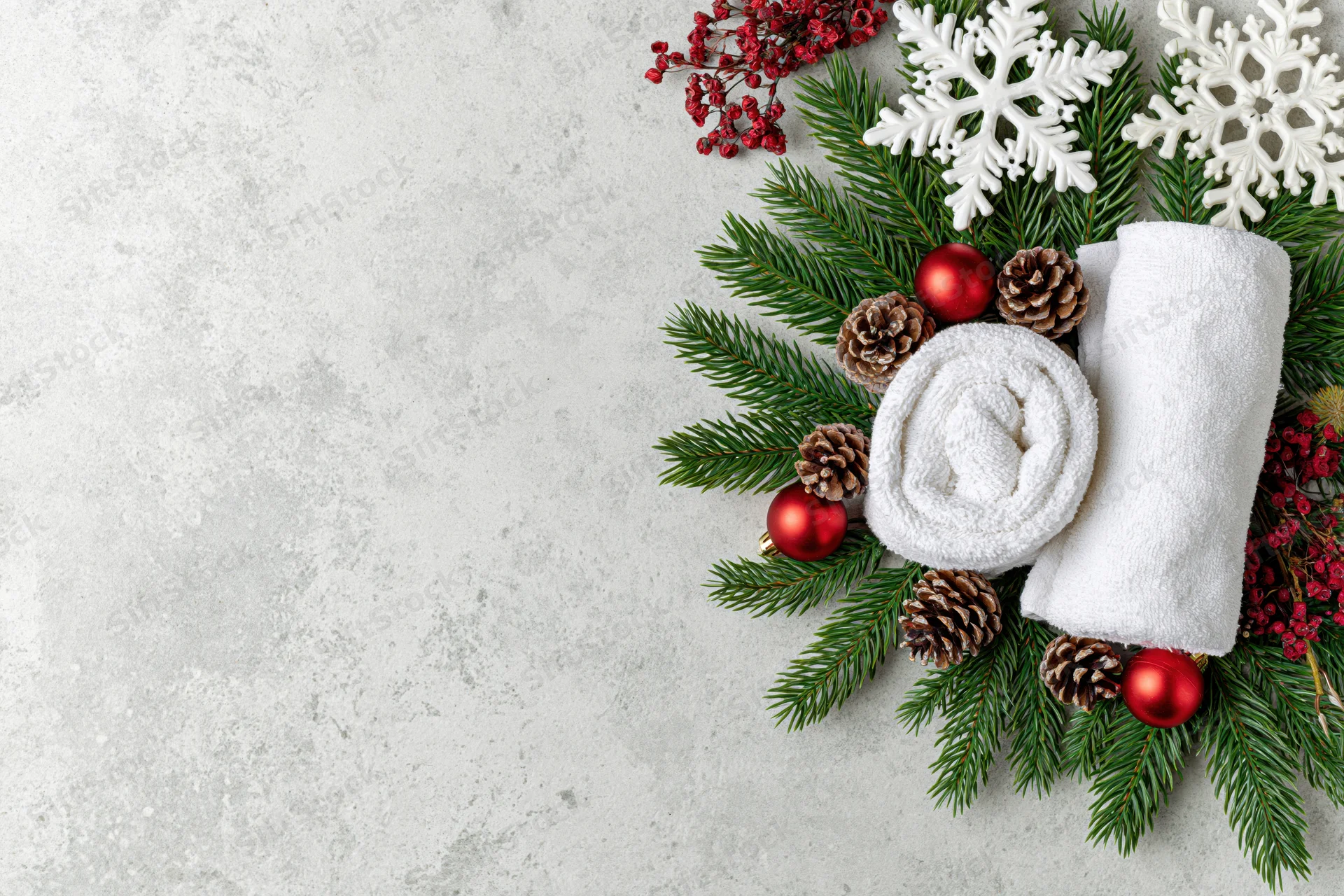 Two rolled white spa towels arranged with evergreen branches, pinecones, red ornaments and snowflakes on a gray concrete surface