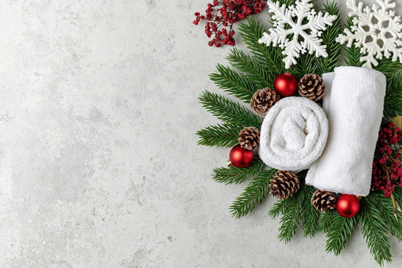 Two rolled white spa towels arranged with evergreen branches, pinecones, red ornaments and snowflakes on a gray concrete surface