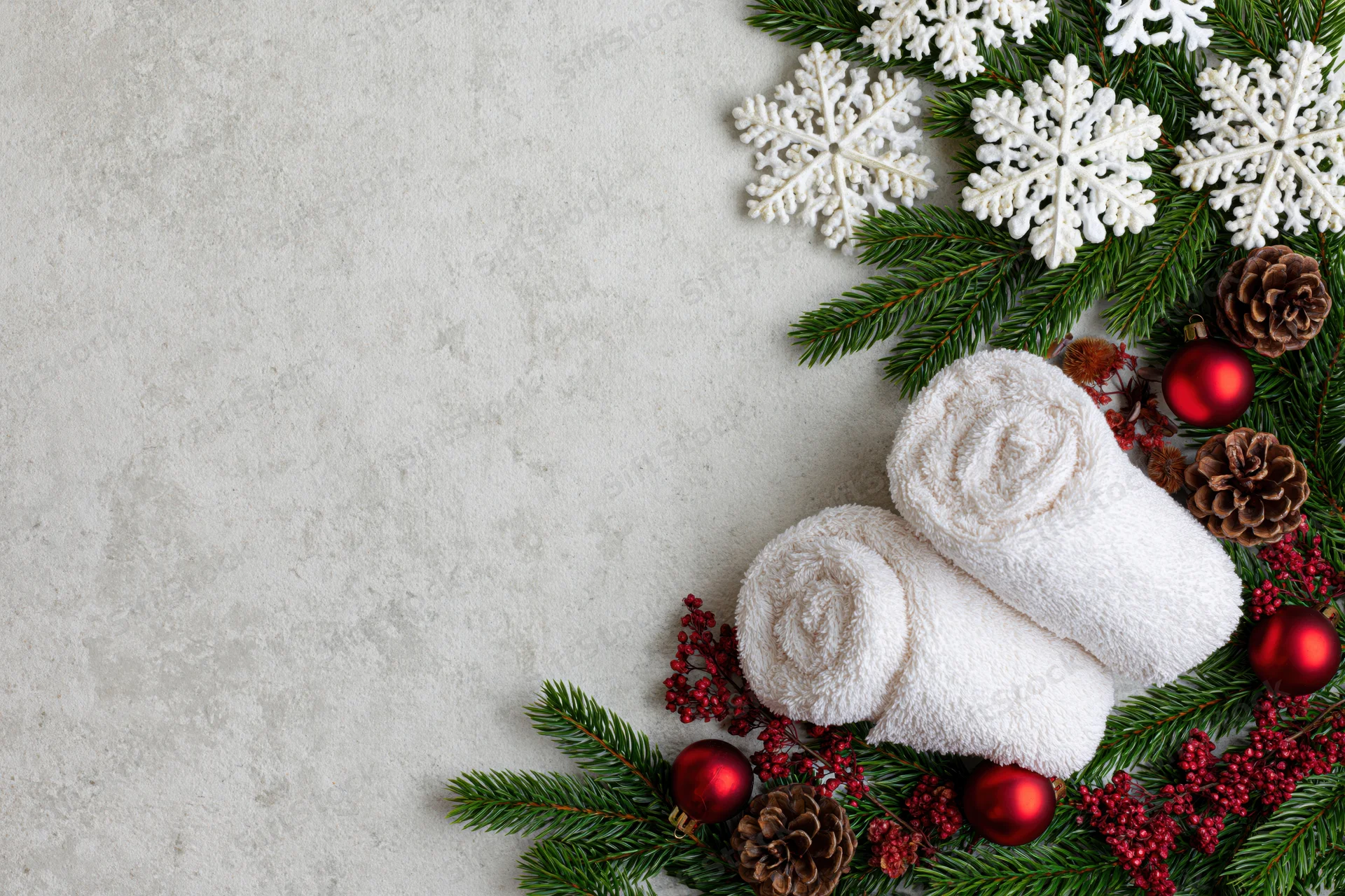 Two rolled white spa towels arranged with evergreen branches, snowflakes, pinecones and red ornaments on a light textured surface