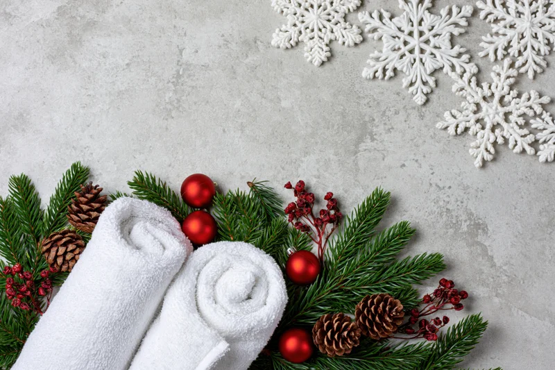 Two rolled white spa towels arranged with pine branches, red ornaments, pinecones and berries on a textured surface for christmas decoration